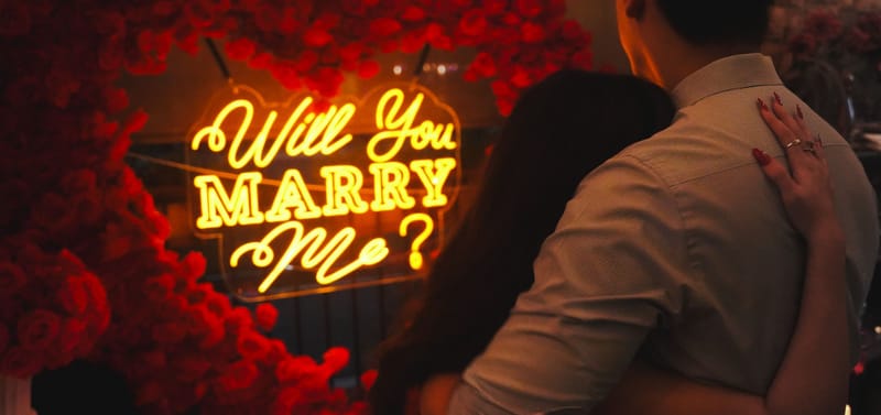 Couple embracing in front of a glowing Will You Marry Me neon sign surrounded by red roses, Phoenix proposal