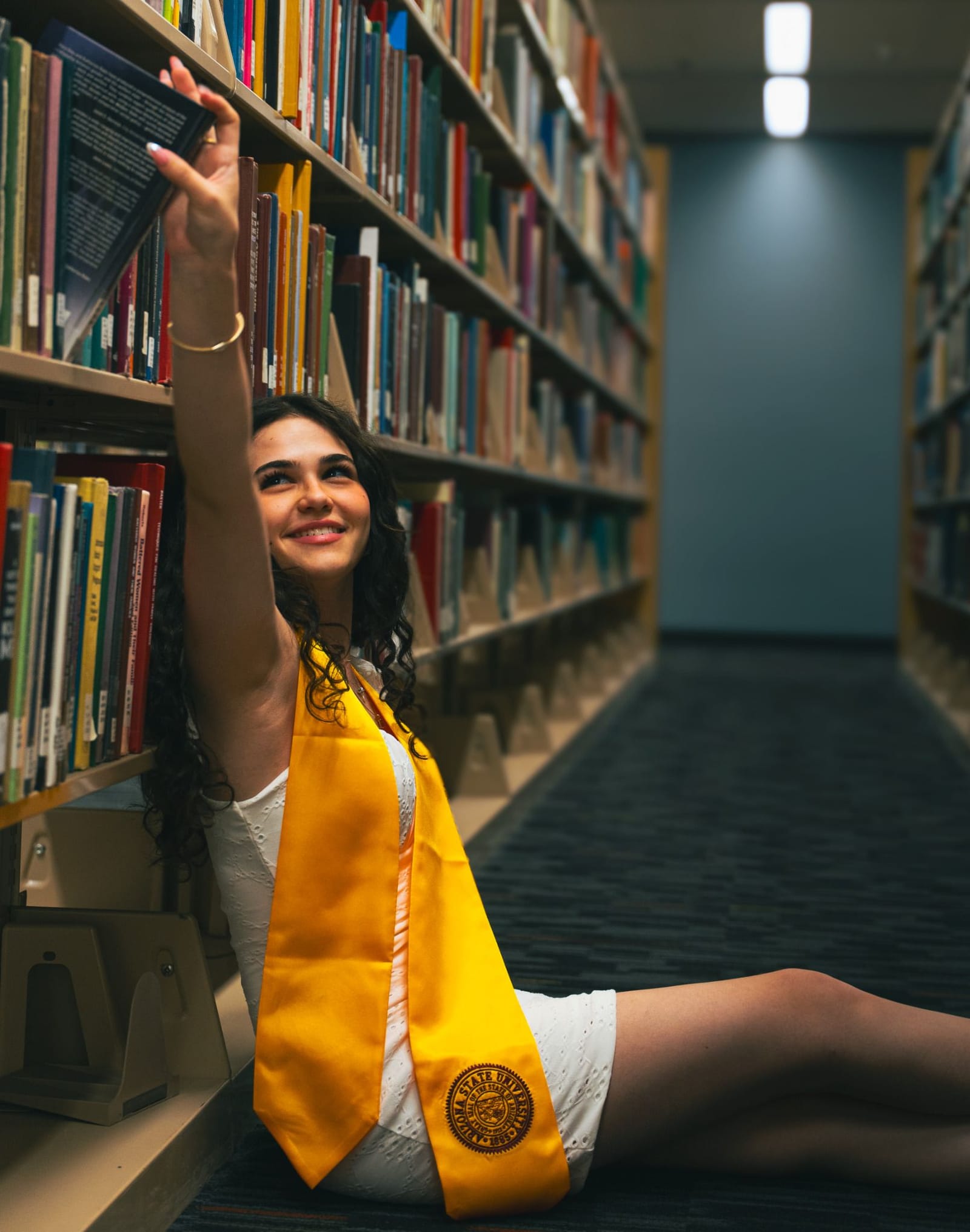 ASU graduate in yellow dress reaching for a book while seated on a stack in the library, Tempe