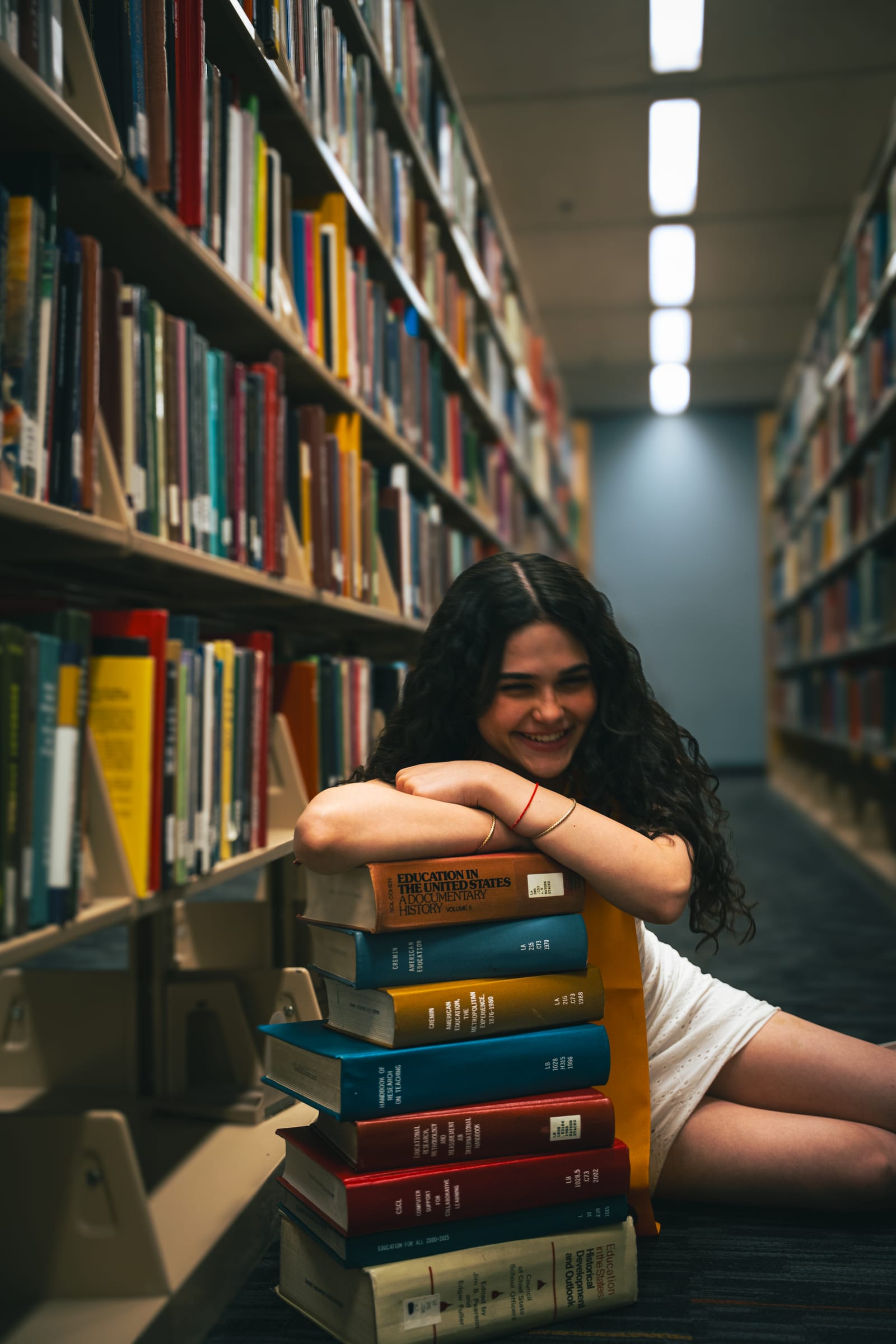 ASU graduate in yellow dress seated on a stack of books in a library aisle, Tempe, Arizona