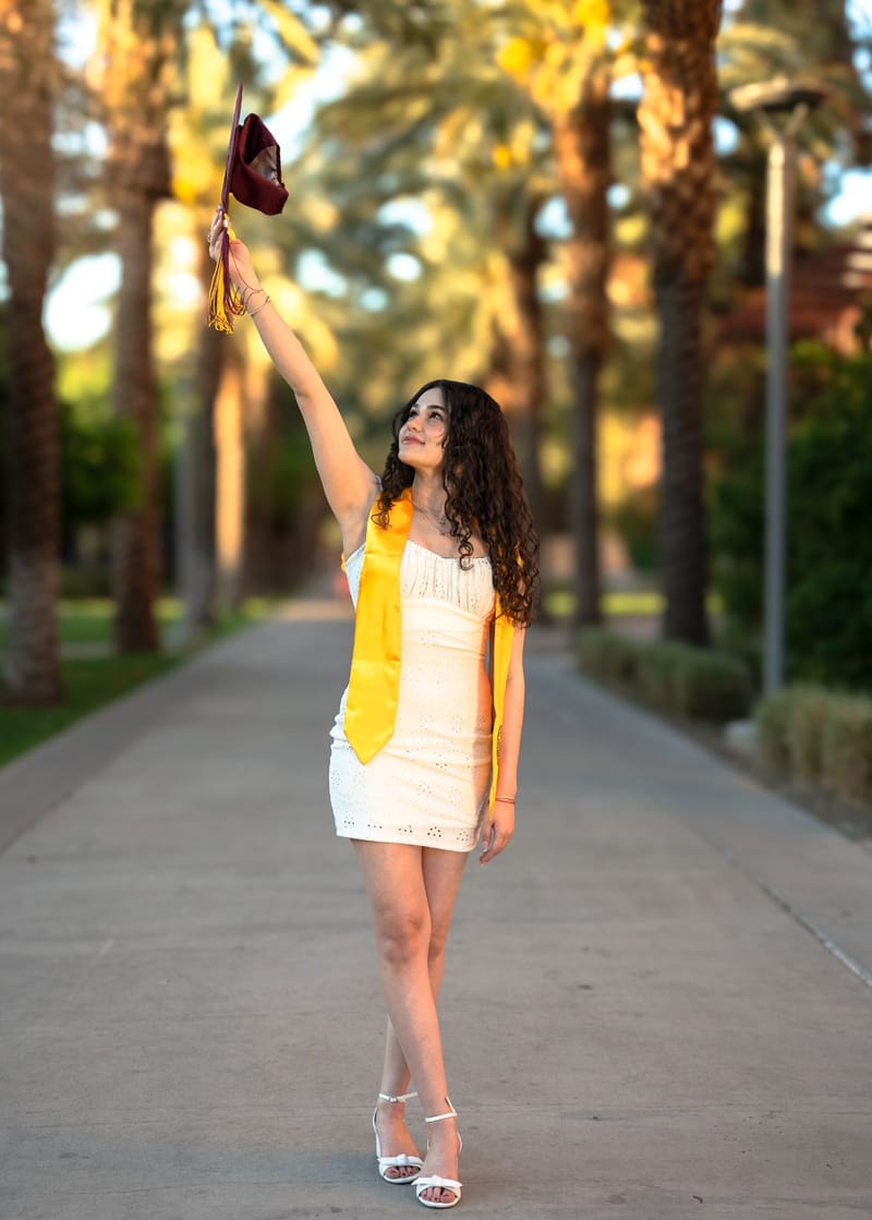 ASU graduate in white dress and gold stole raising her cap on Palm Walk at golden hour, Tempe