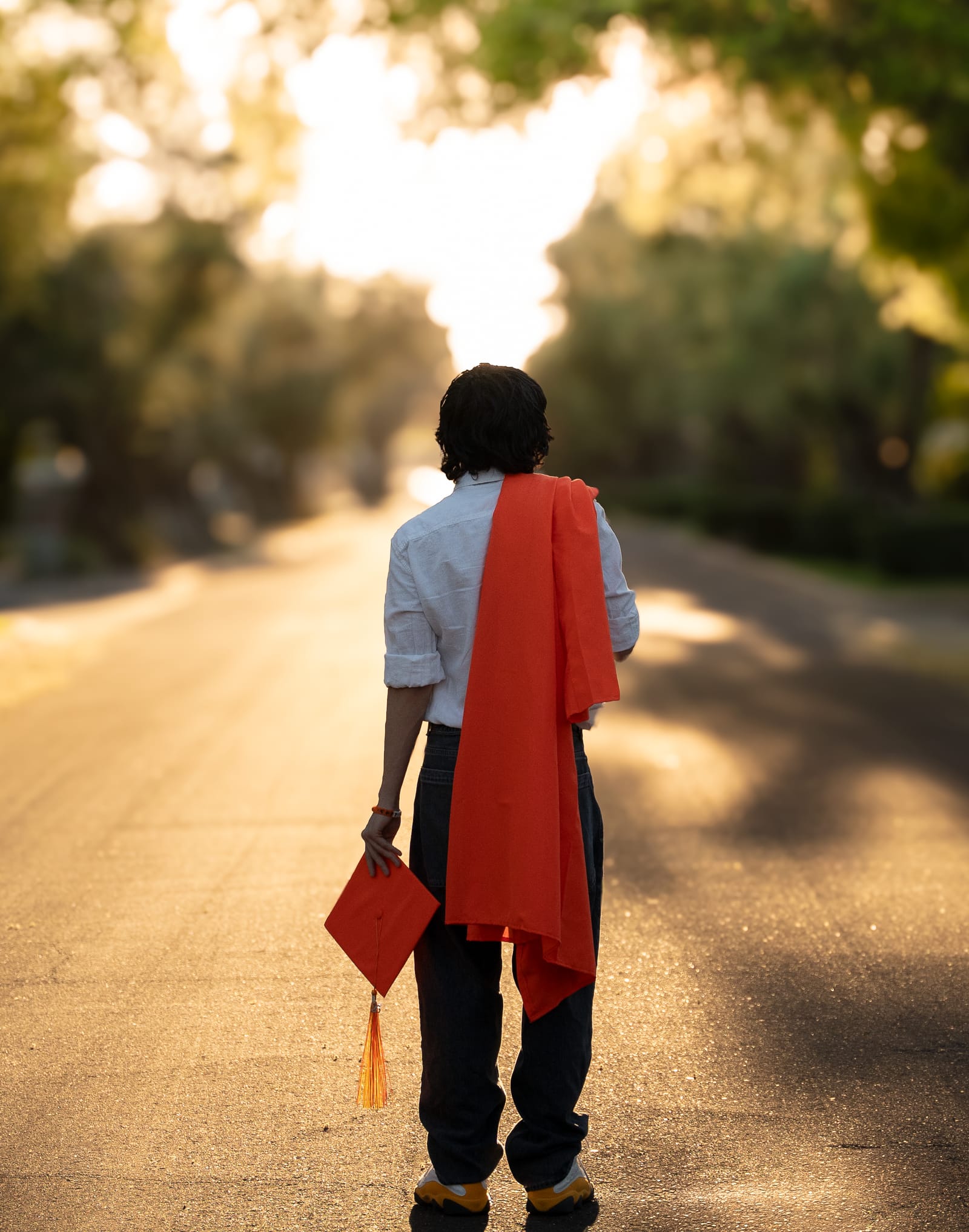 Graduate walking down a backlit neighborhood road carrying his cap, Phoenix, Arizona