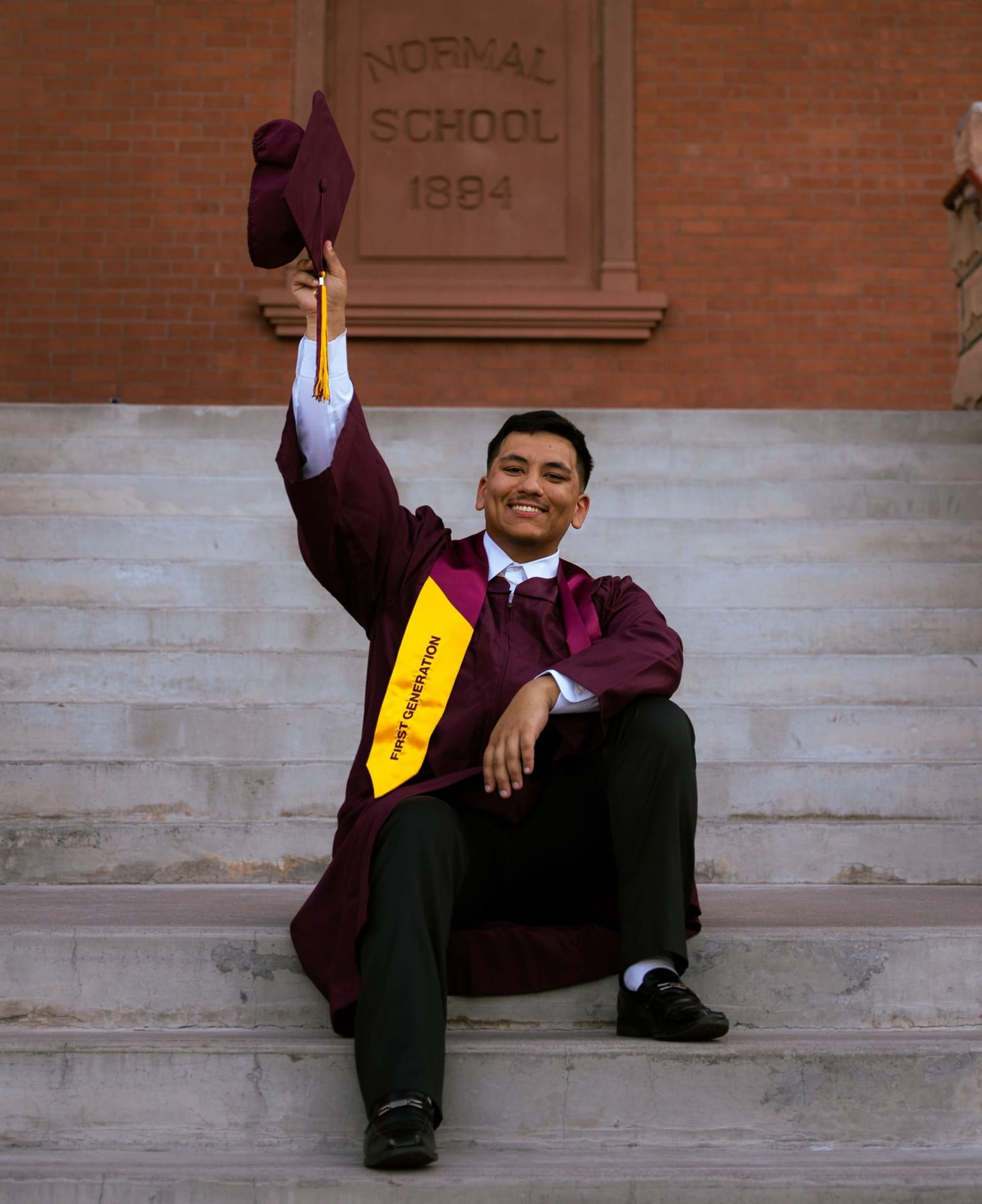 First generation ASU graduate raising cap on campus steps in maroon and gold, Tempe, Arizona