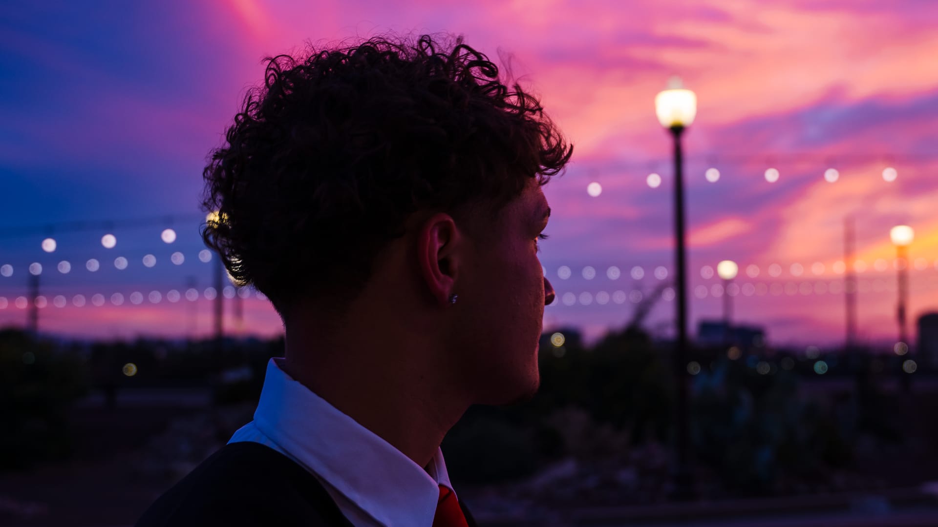 High school graduate in gown and red tie at dusk under string lights, Phoenix, Arizona
