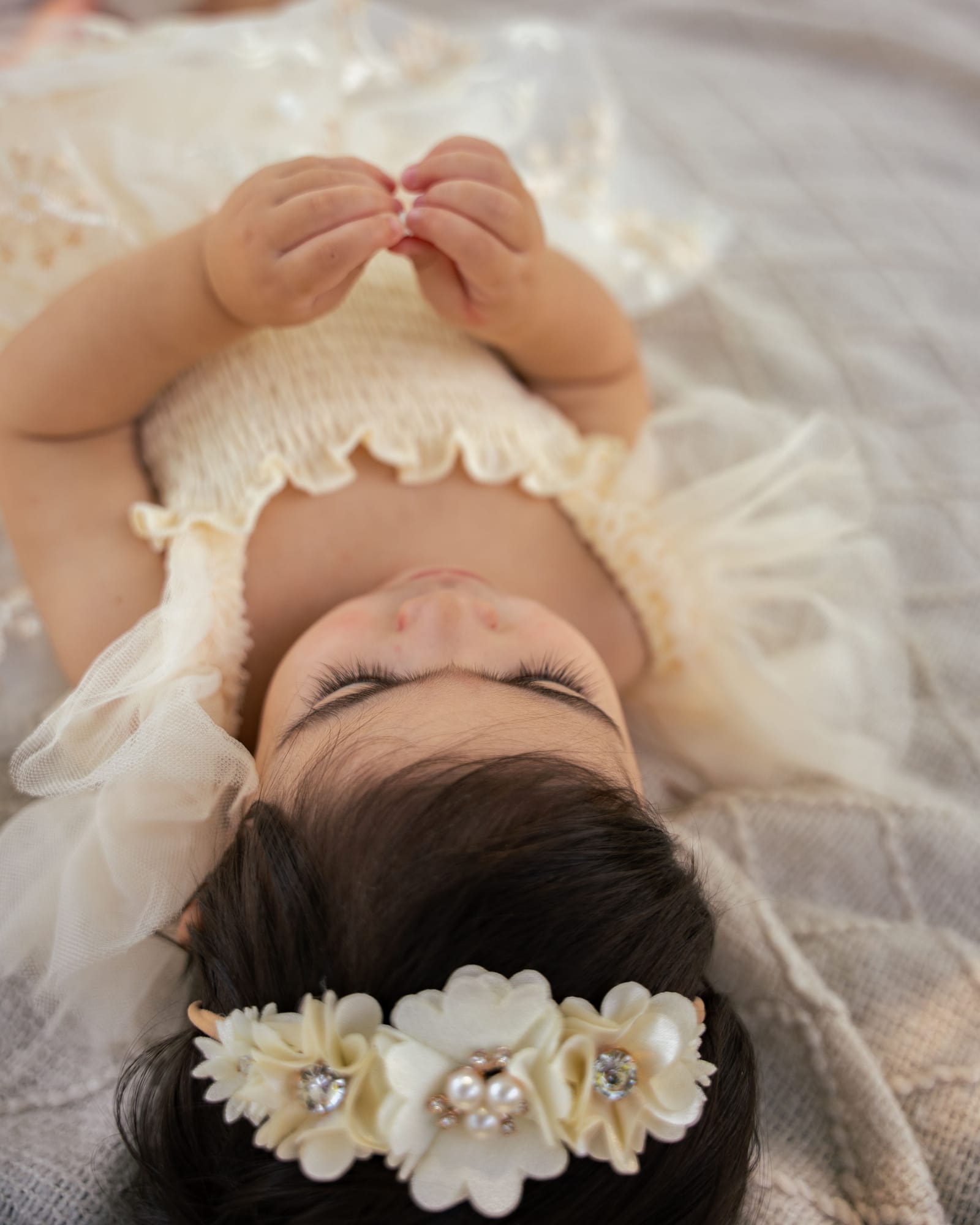 Overhead photo of a baby in a cream dress and pearl flower headband lying on a knit blanket, Roslyn, New York
