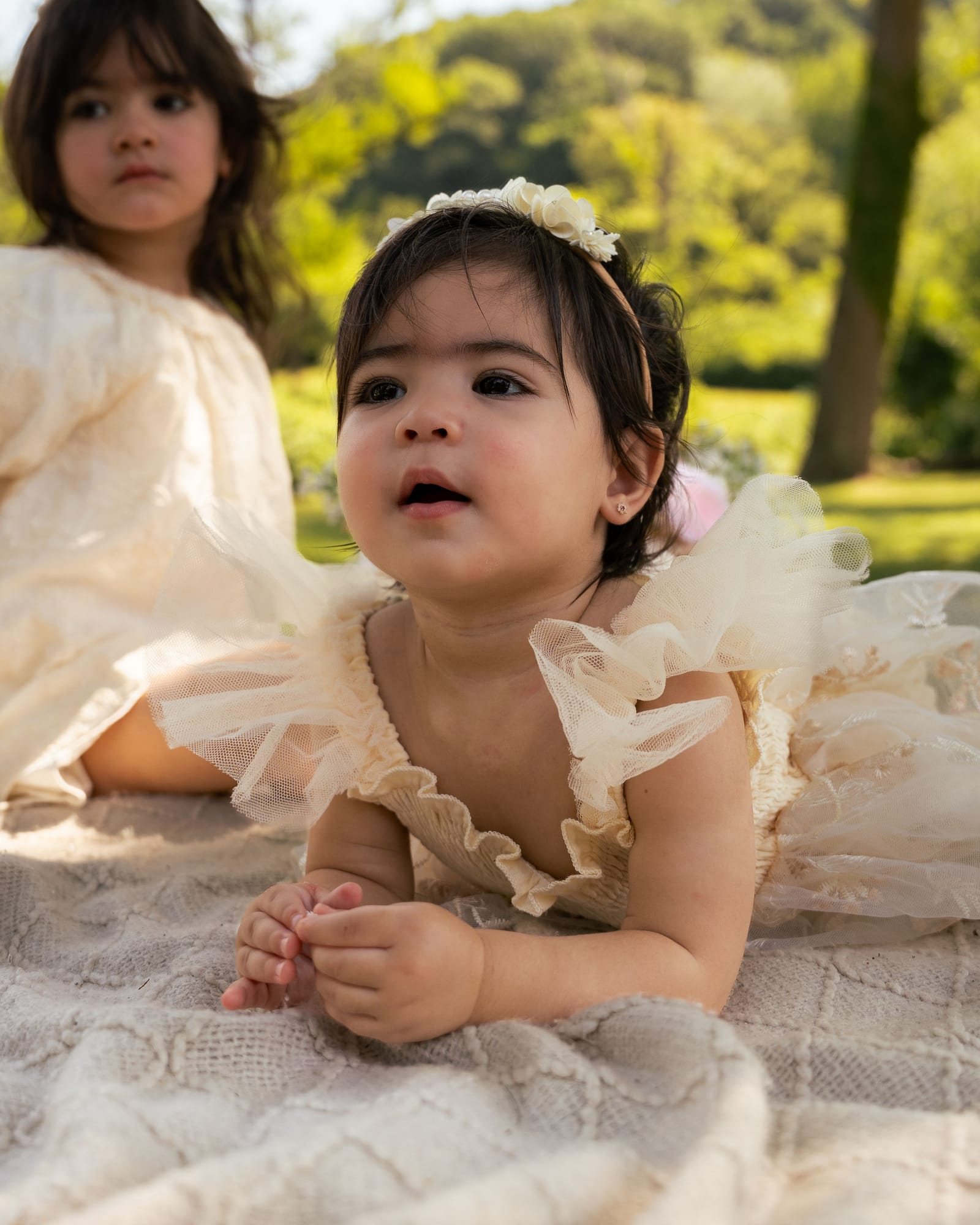 Baby in a tulle dress lying on a blanket looking up, her sister behind her in the grass, Roslyn, New York