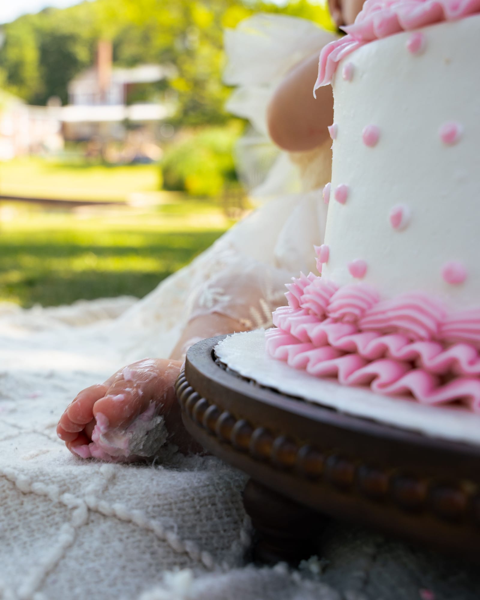 Close detail of a baby hand covered in frosting beside a pink ruffled first birthday cake, Roslyn, New York