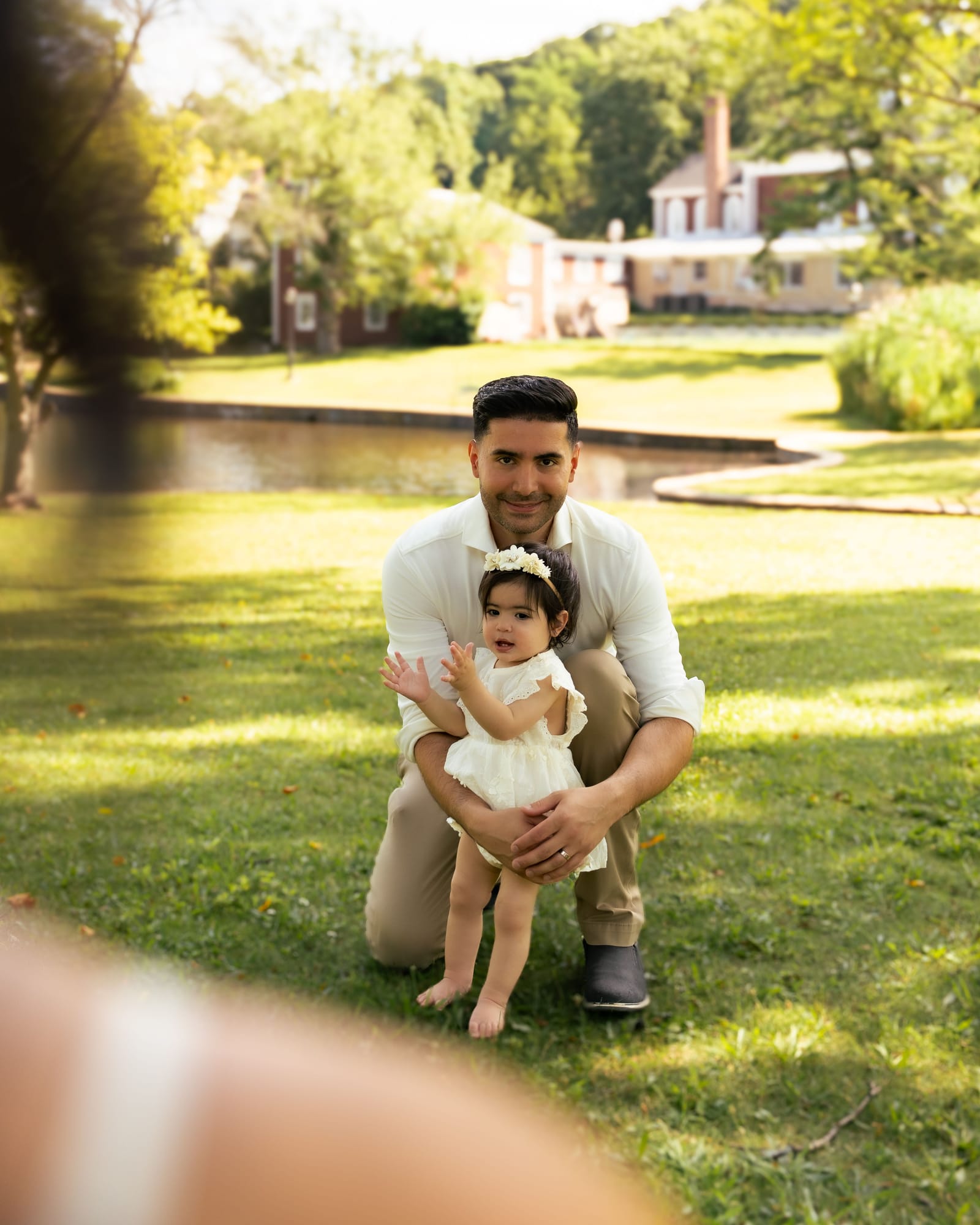 Father kneeling on the grass holding his baby daughter in front of a pond, Roslyn, New York