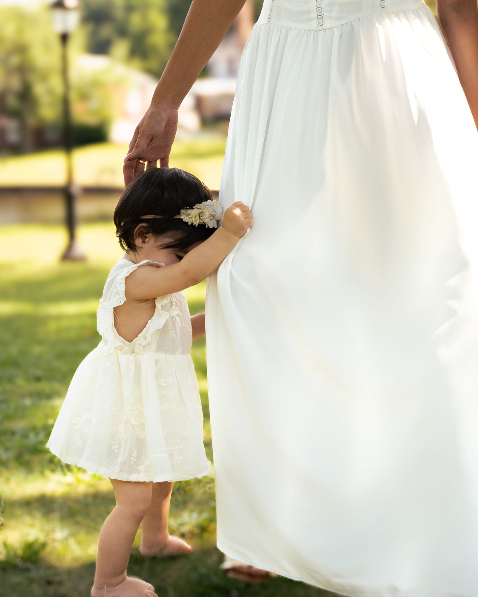 Toddler in a lace dress holding her mother hand and hiding in the hem of her white gown, Roslyn, New York