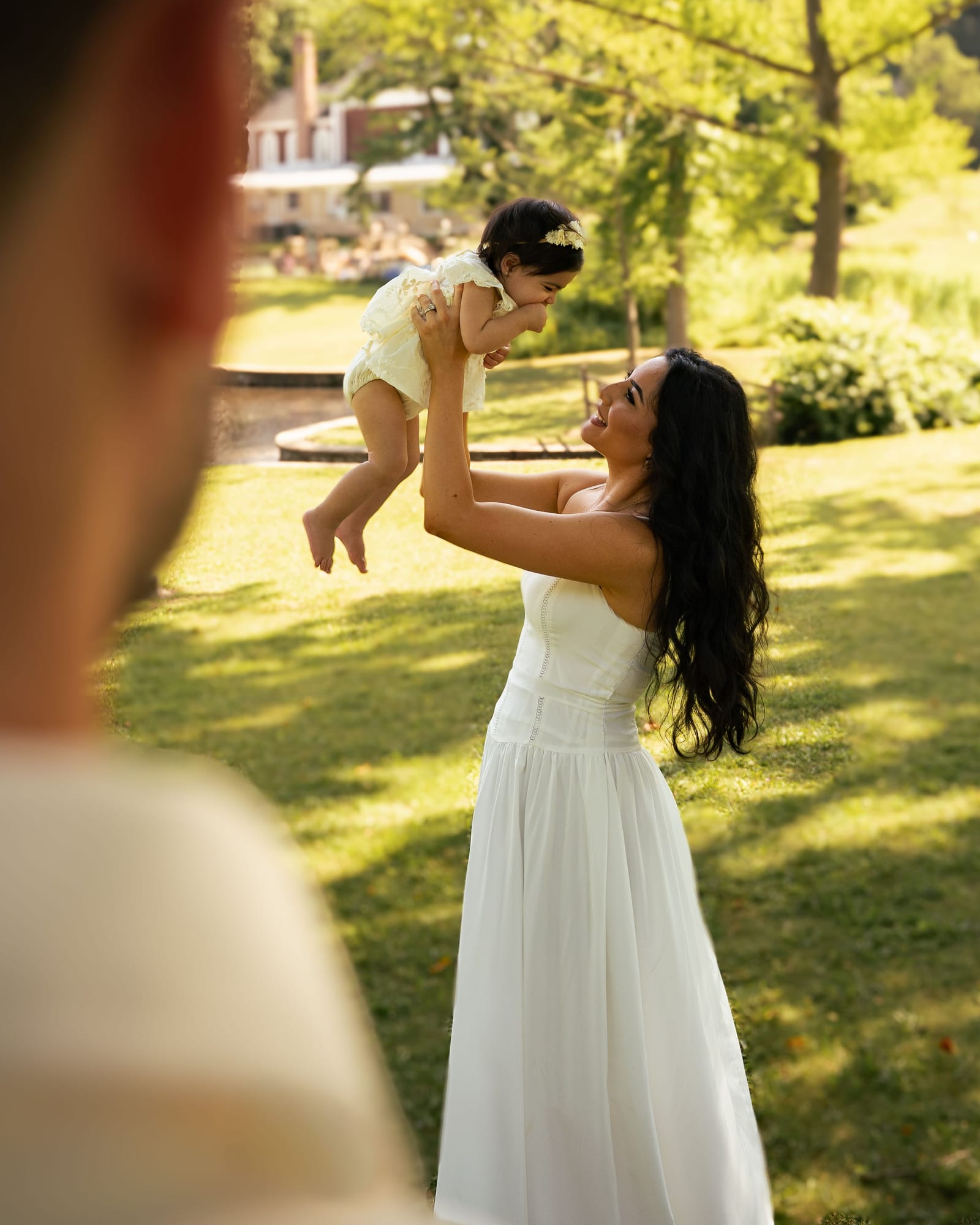 Mother in a white dress lifting her daughter into the sunlight, seen past the father shoulder, Roslyn, New York