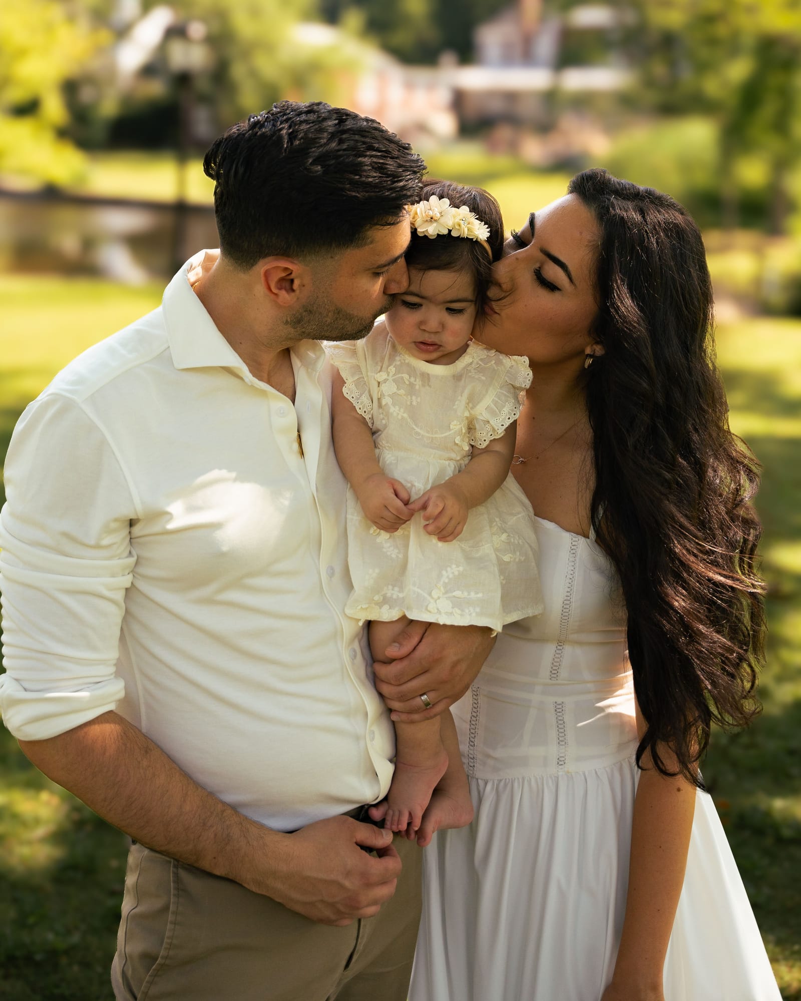 Parents in cream holding and kissing their one year old daughter in a sunlit park, Roslyn, New York